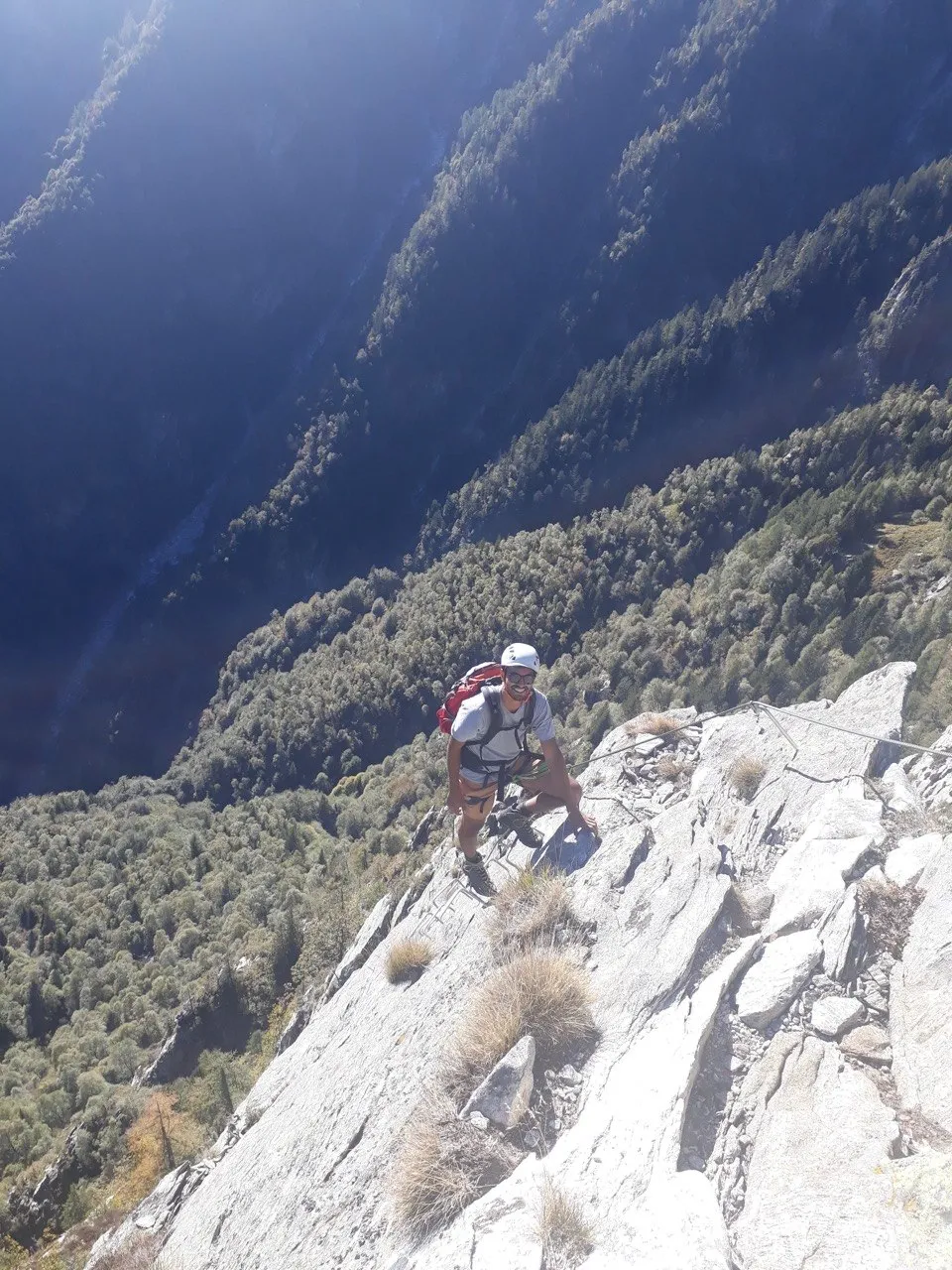 A climber crouches on a pale rock ridge that drops steeply into a deep, forested valley.