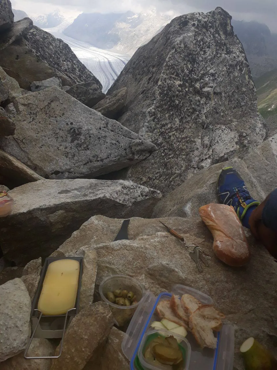 A raclette break among the rocks: melted cheese in a pan on a small stove, with bread, olives and pickles, and the Aletsch Glacier visible through a gap in the rocks behind.