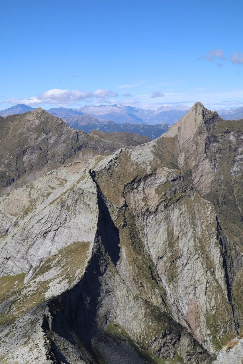 Jagged grey-green rock ridges and a deep couloir drop away toward hazy ranges under a clear blue sky.