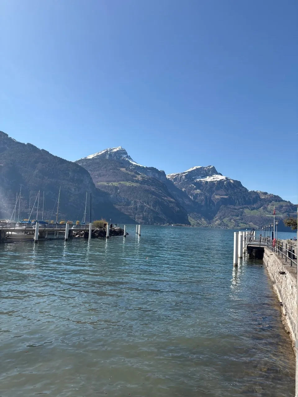 A small boat dock with sailboat masts, snow-capped mountains rising across the lake.