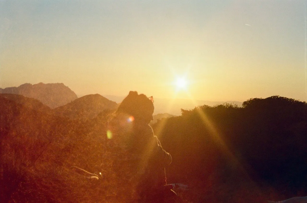 A hiker silhouetted against a bright sun setting behind layered mountain ridges.