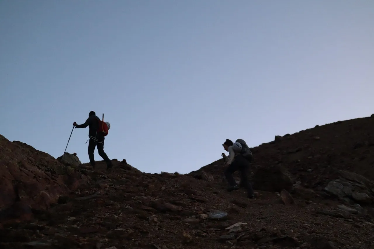 Two hikers silhouetted as they climb reddish scree in the early dawn light.