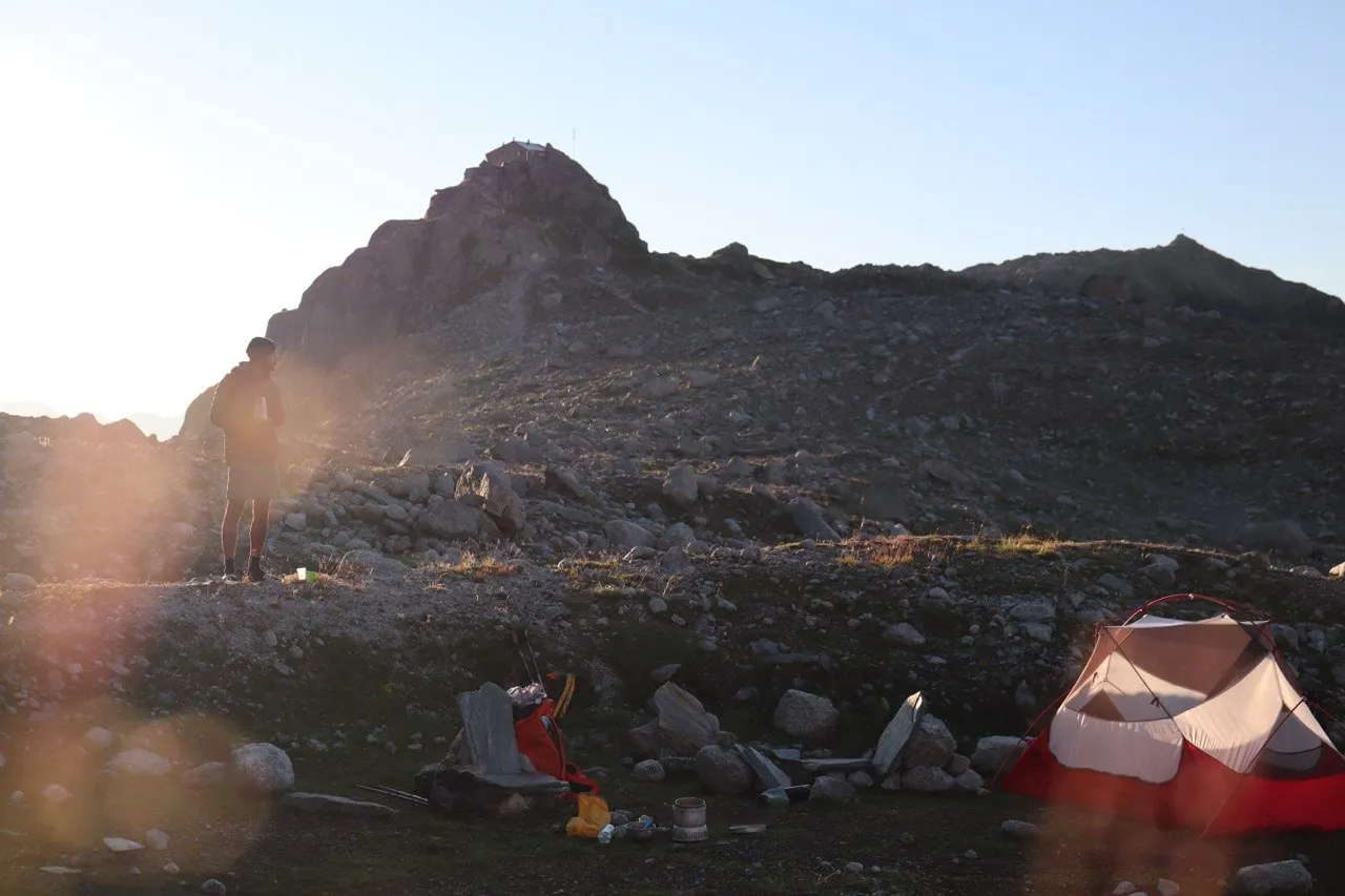 Dawn at a rocky bivouac camp with a red tent, scattered gear, a hiker standing, and the summit hut silhouetted on the peak behind.