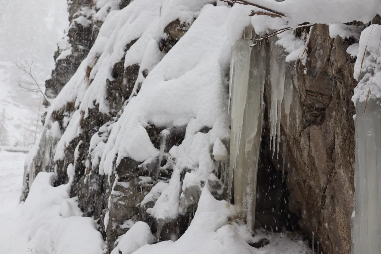 A rock face draped in snow and long blue-white icicles during a snowfall.