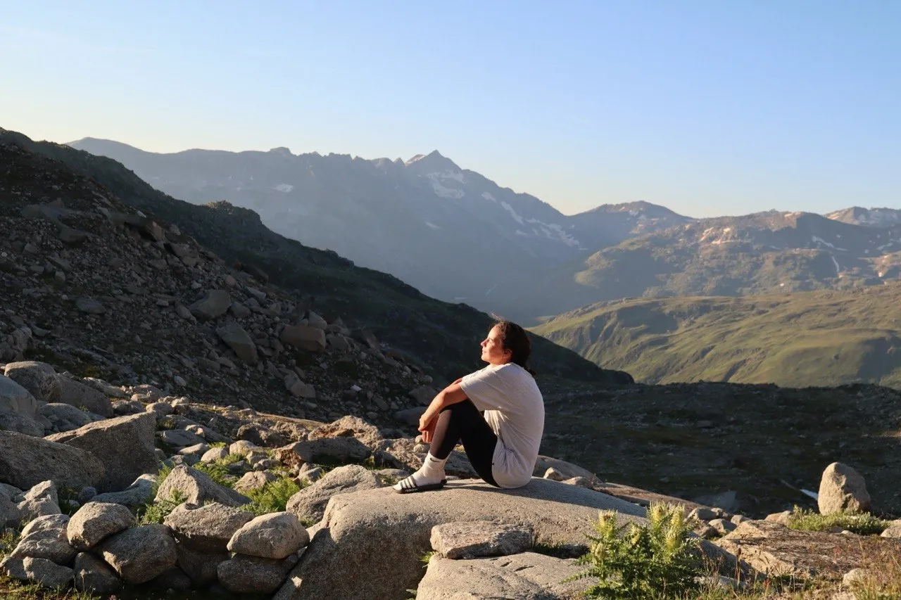 Una excursionista sentada sobre un bloque de granito bajo la luz dorada del atardecer, contemplando las brumosas cadenas montañosas.