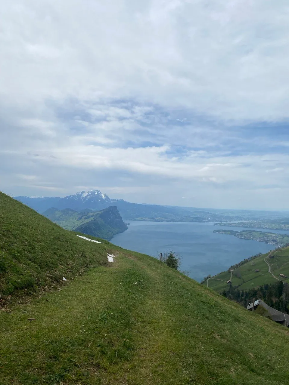 A grassy ridge path with patches of snow leads down toward the lake, a distinctive peak rising in the background.