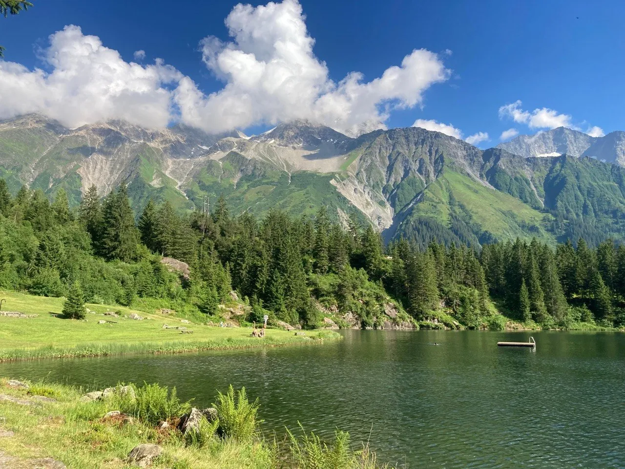 The turquoise-green Golzerensee sits nestled between forest and rugged mountain peaks, with a small jetty reaching into the calm water.