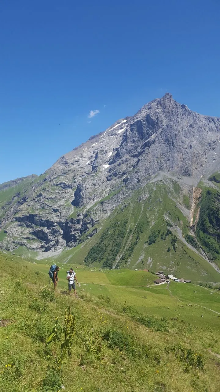 Two hikers climb a steep alpine meadow below a pyramid-shaped rock peak, with a few alp huts in the middle distance.