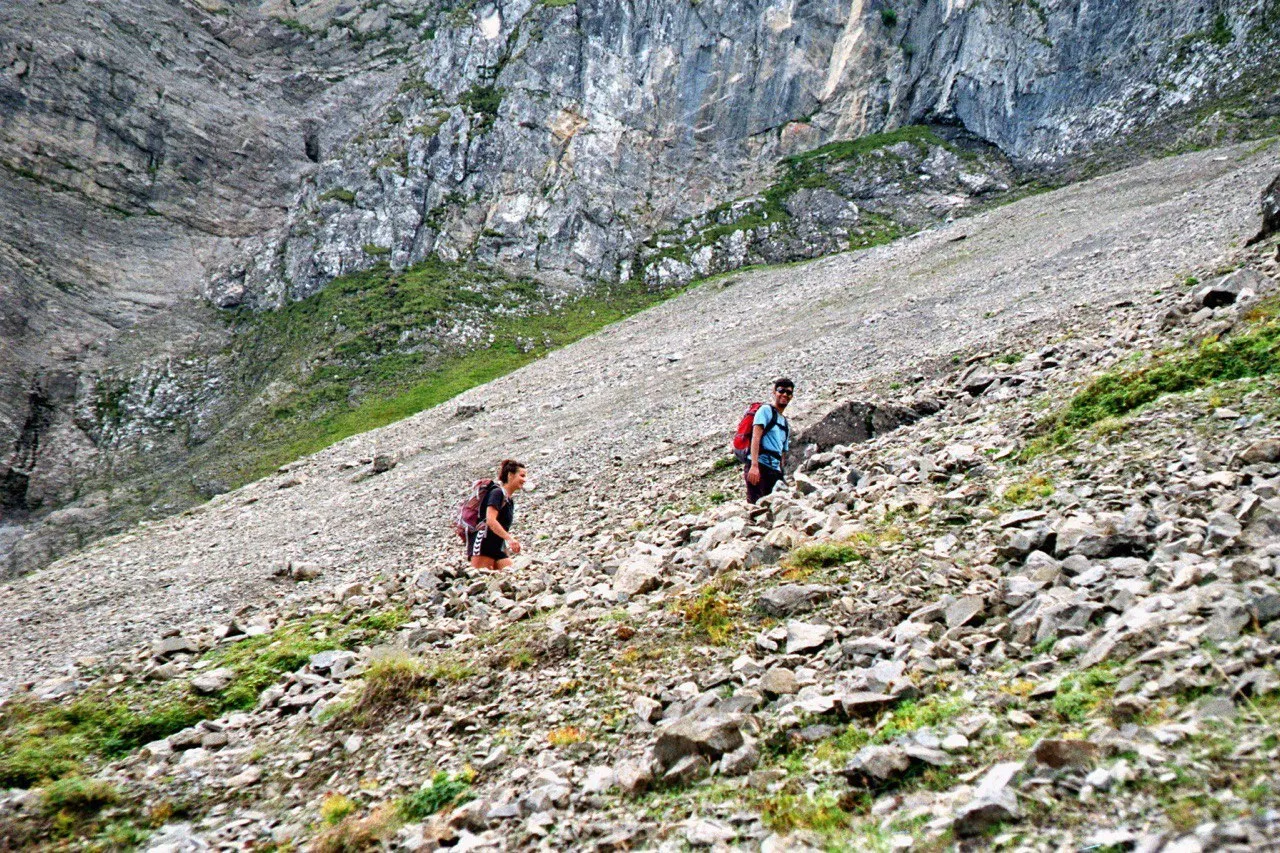 Two hikers climbing a broad scree slope beneath grey limestone cliffs.