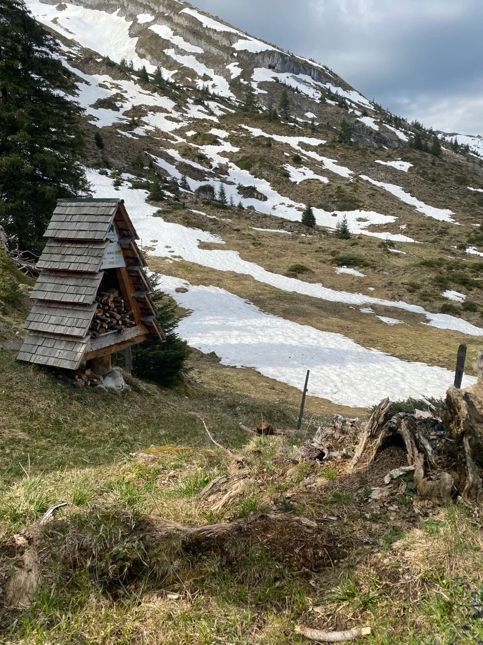 A small wooden shelter with a shingled roof on the hillside, surrounded by thawing grass and lingering patches of snow.