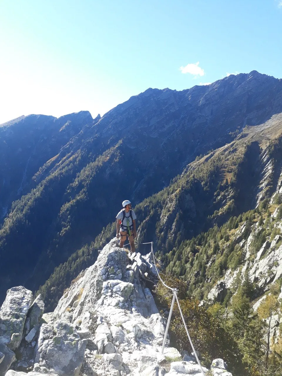 A helmeted climber stands on a narrow rock spur high above a forested valley, a massive, rocky mountain ridge opposite under a blue sky.