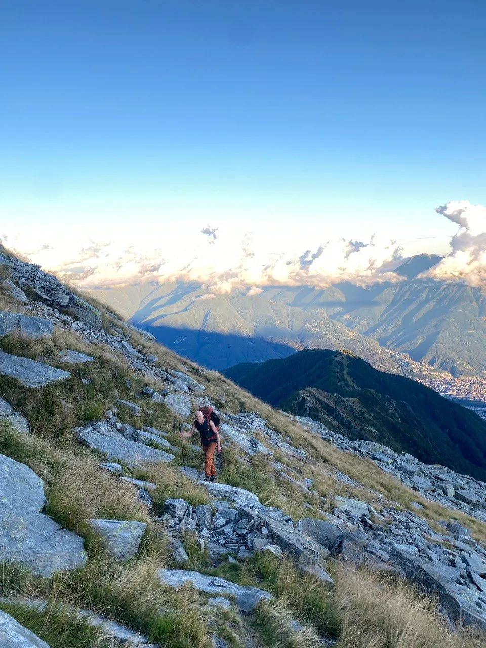 A hiker with a red backpack climbs a grassy, rocky slope, a sea of clouds filling the valley behind and a town glimpsed far below.