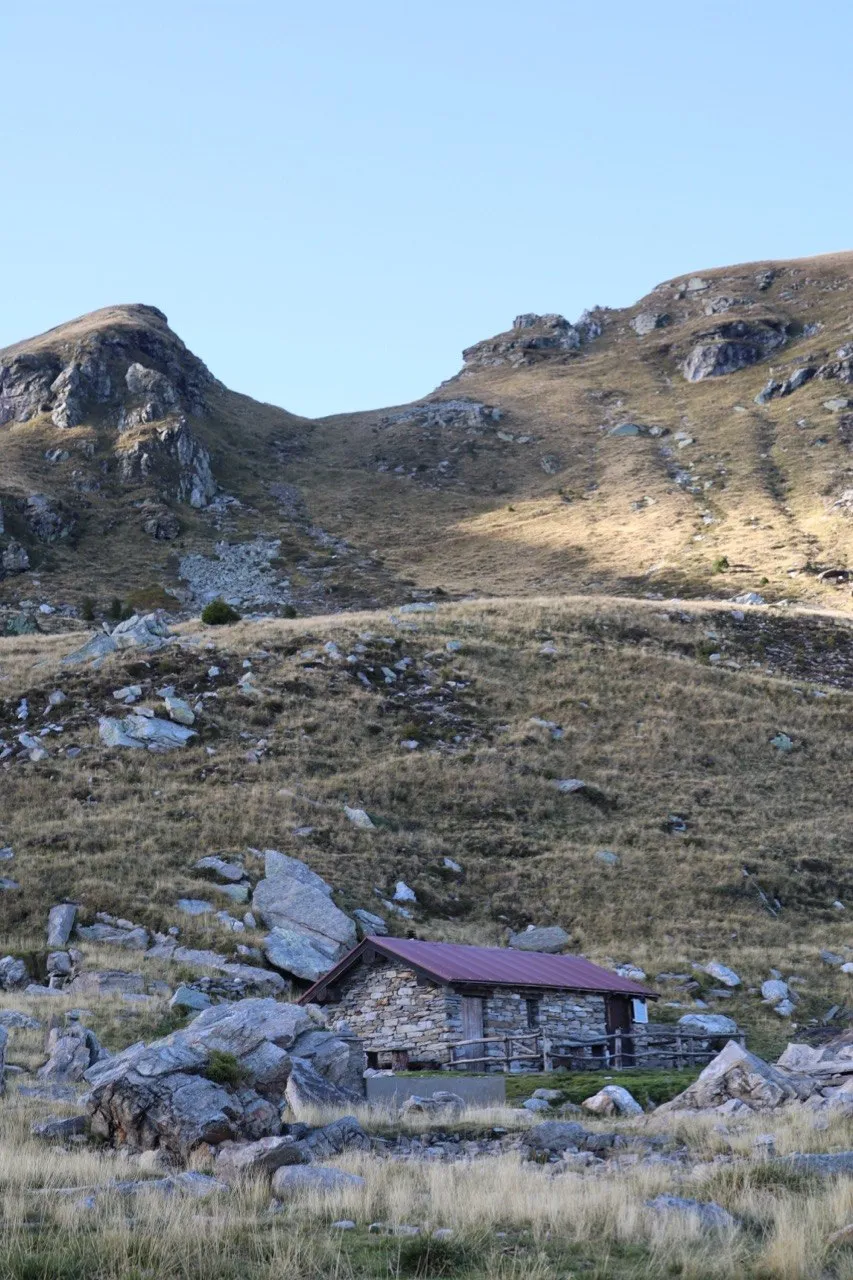 A stone hut with a red roof tucked beneath grassy slopes and rocky knolls in the early light.