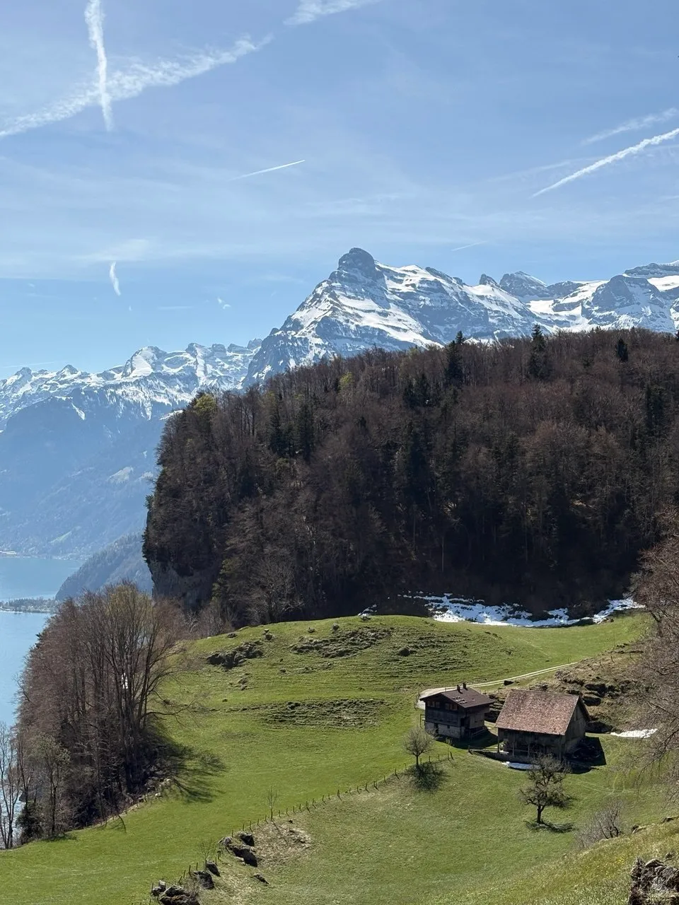 An old hay barn and farmhouse on a steep green meadow high above the lake, with forested slopes and snow-capped peaks behind.