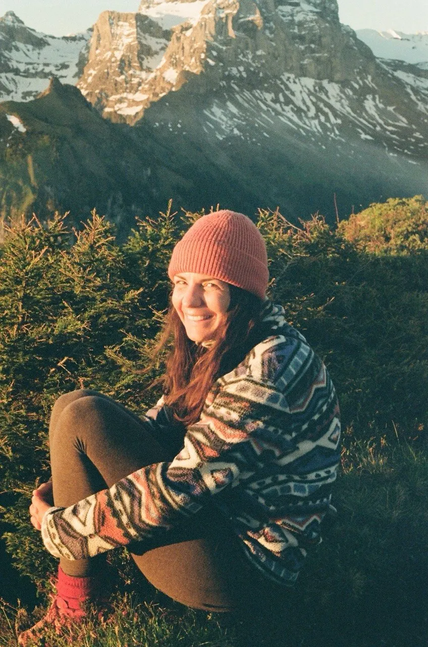 A hiker in a pink beanie and patterned fleece sits on a grassy slope in the evening light, jagged snow-streaked peaks rising behind.