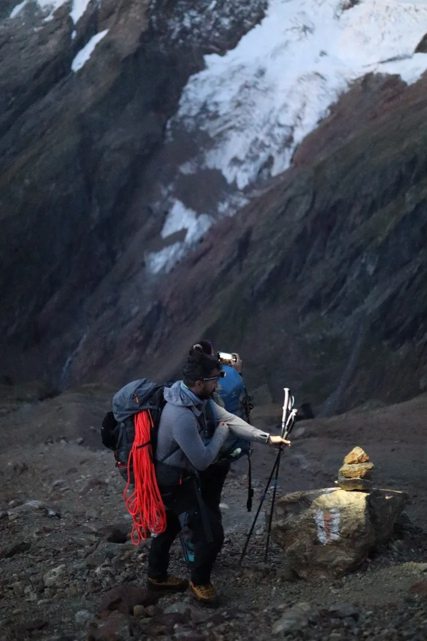 Hiker with a coiled red rope on his pack beside a marked cairn.