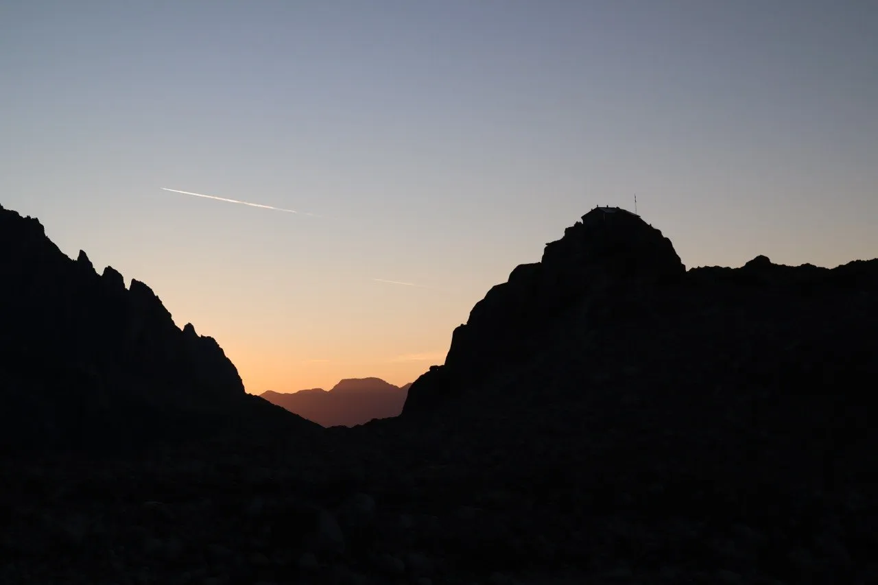 Mountain silhouettes at dusk with a hut perched on a ridge and an orange afterglow on the horizon.