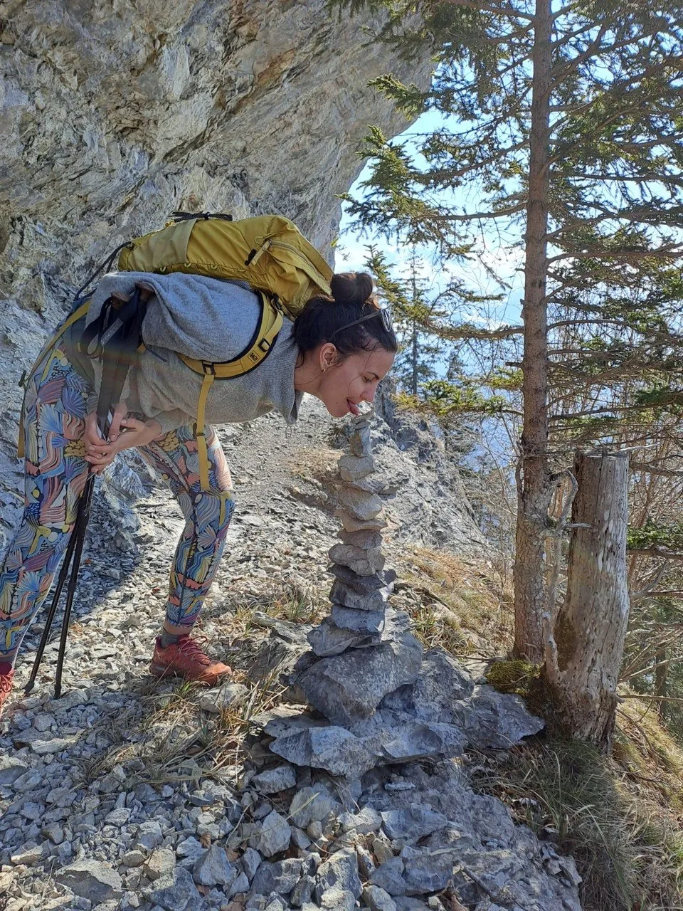 A hiker with a yellow backpack bends to inspect a small stone cairn on a rocky ridge.