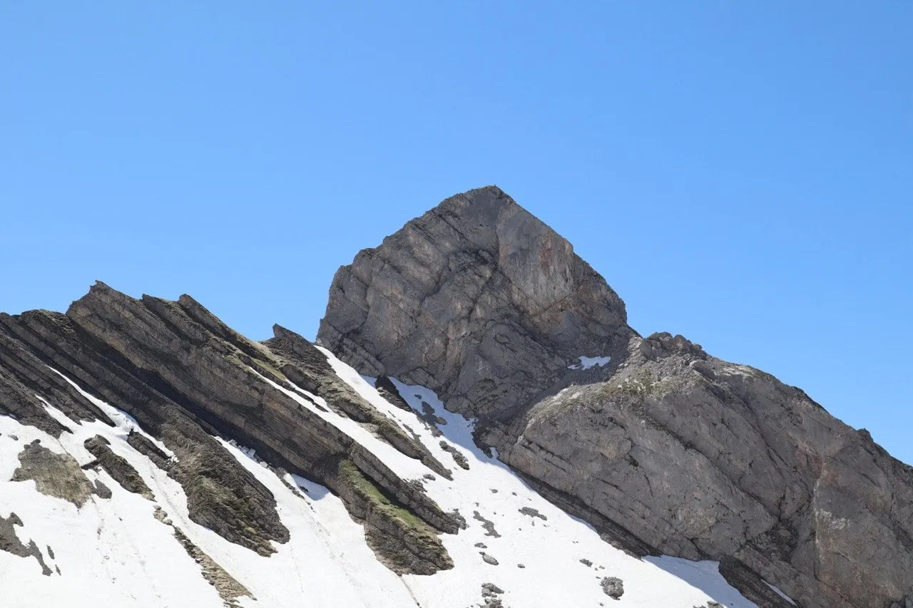 Layered rock folds streaked with snow lead up to a rugged summit block.