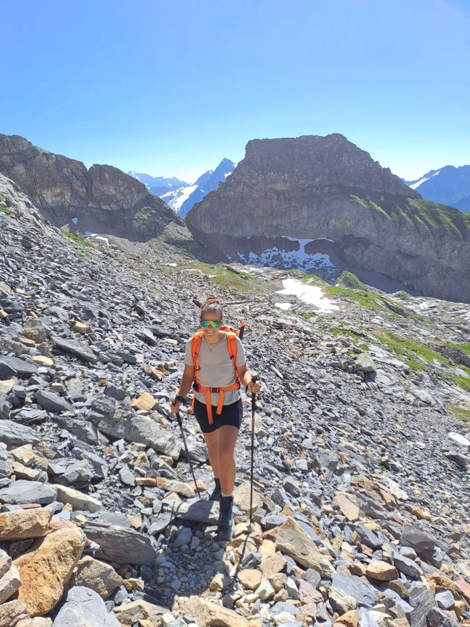 A hiker with trekking poles crosses a scree field, a striking rock tower with patches of snow behind her.