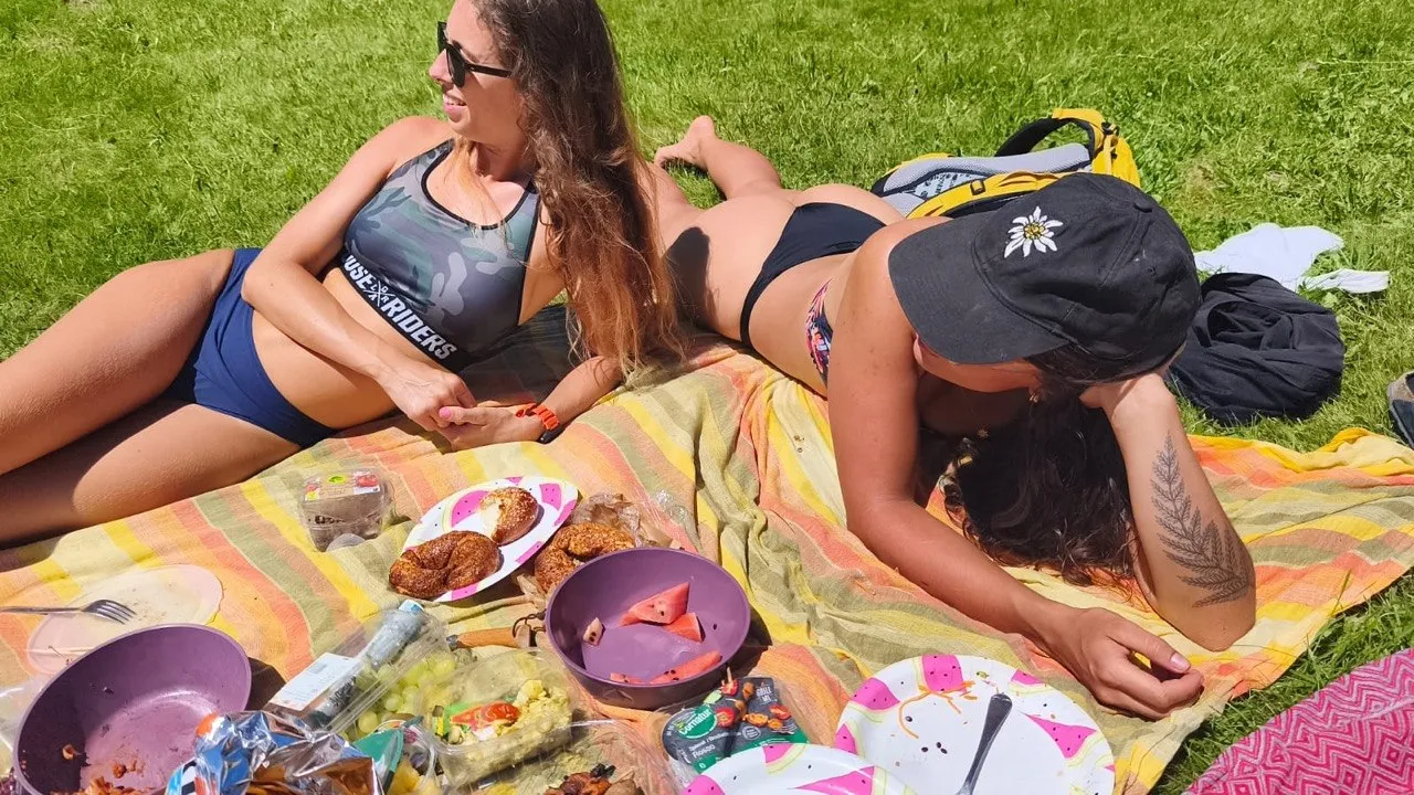 Two women relax on a picnic blanket in the grass, surrounded by wraps, watermelon and other treats.