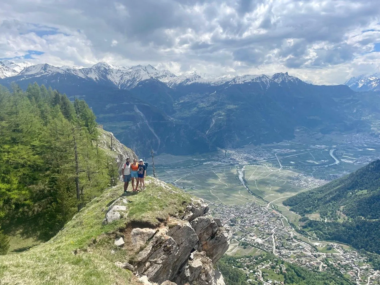 Three hikers pose on a rocky, larch-covered ridge high above the wide Rhone valley, snow-capped peaks and a patchwork of fields far below.