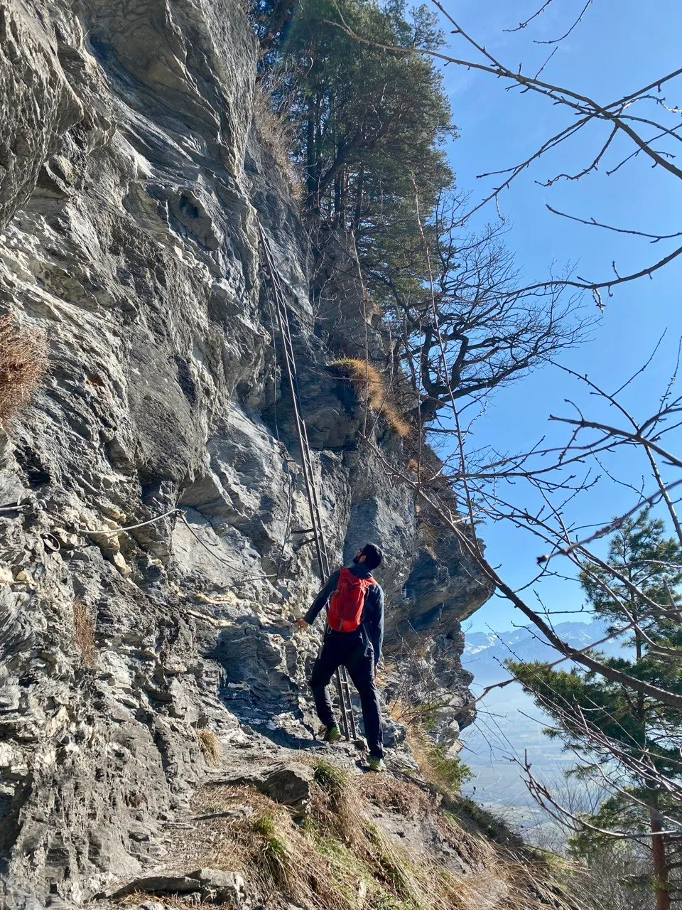 A climber holds onto a fixed rope on a steep rock face, with a wide valley view behind.
