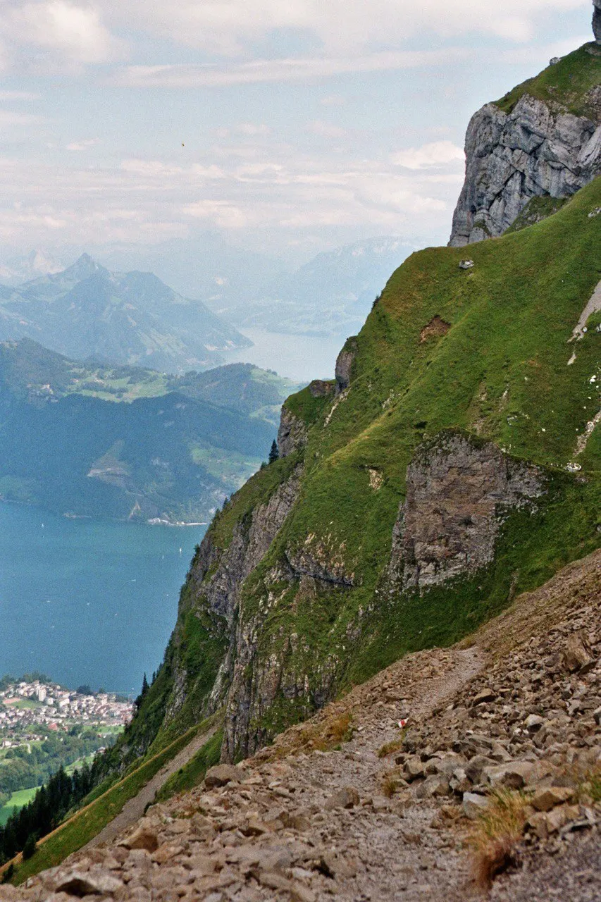 A narrow path traversing a steep green flank high above Lake Lucerne, hazy peaks beyond.