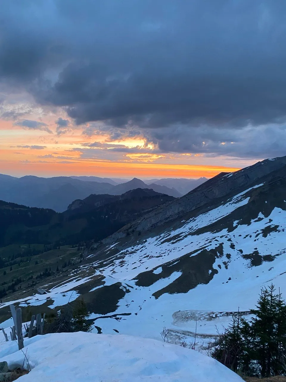 Dawn over a snowy slope, the sky glowing orange on the horizon above layered mountain ranges.