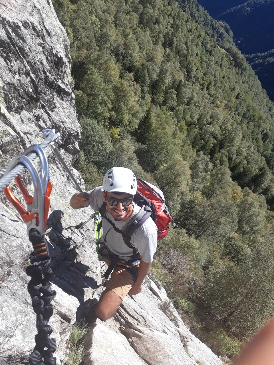 A climber looks up at the camera with a grin, carabiners and a via ferrata set on the steel cable in the foreground, a forested valley behind.