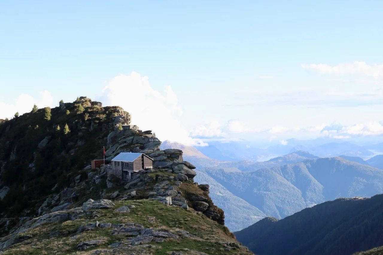 A small wooden mountain hut perched on a rocky ridge, with hazy ridgelines and Lake Maggiore stretching far below under a bright sky.