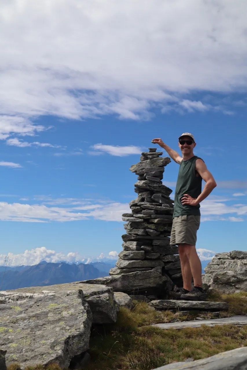 Hiker posing beside a tall cairn on a broad rocky summit under a blue sky.