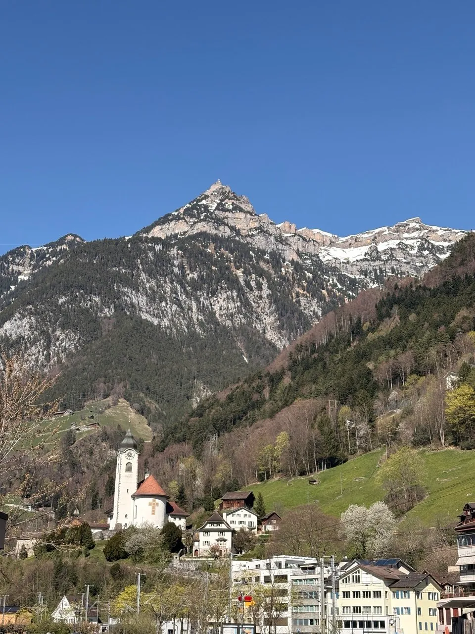 A village with a white church tower below a steep rocky mountain, green spring slopes in between.