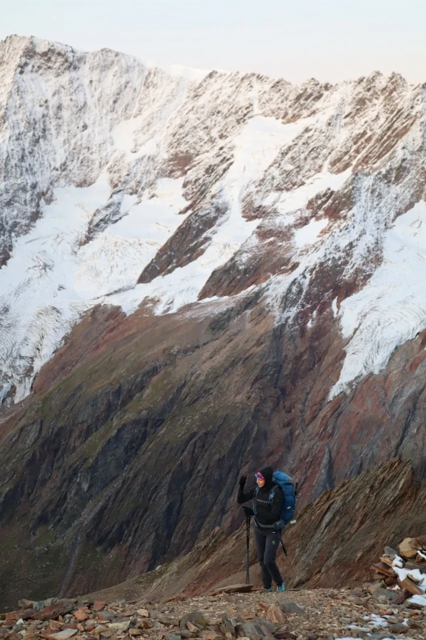 Hiker on a rocky shoulder in front of a big snow-covered mountain face.