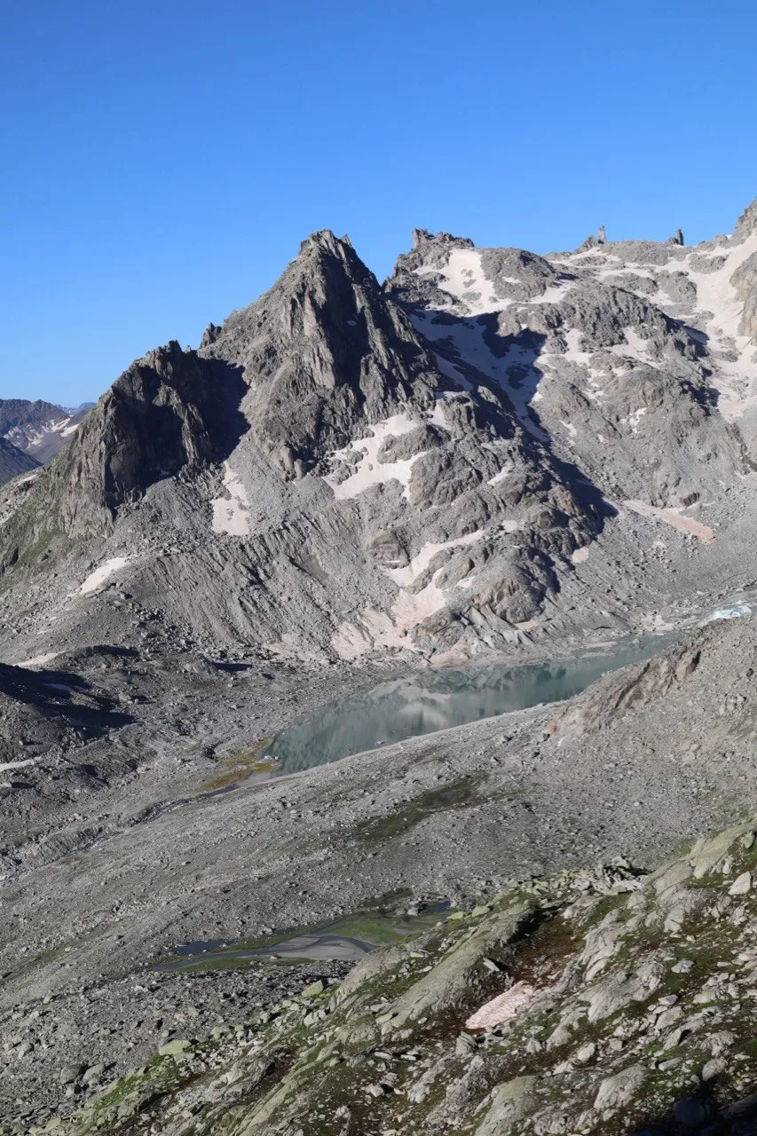 A craggy grey peak streaked with snow above a small turquoise tarn in a rocky basin, under a clear blue sky.