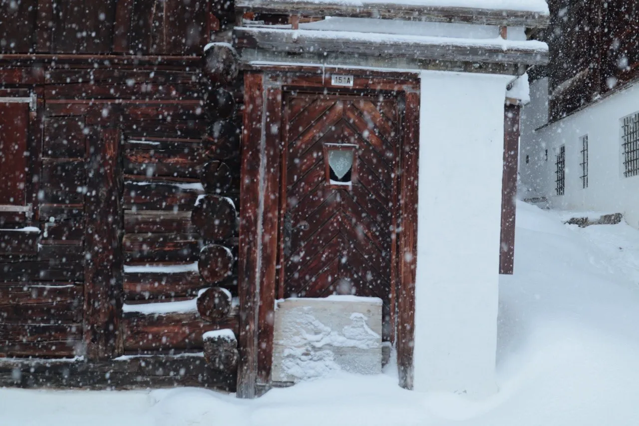 A dark timber chalet's carved wooden door, numbered 151A and topped with a small heart-shaped window, framed by whitewashed walls and deep snow.