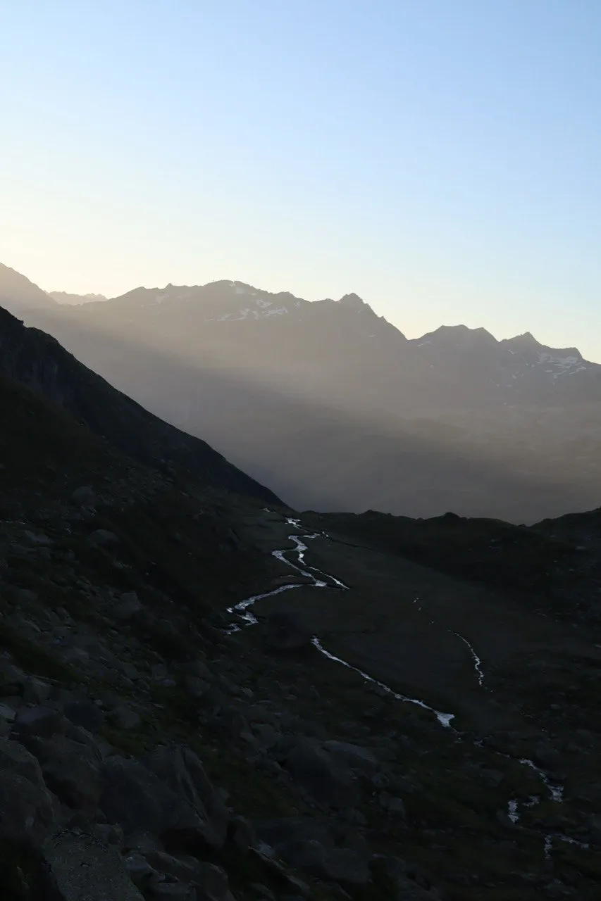 A meltwater stream winds through a high valley at dawn, with hazy ridges receding behind it.