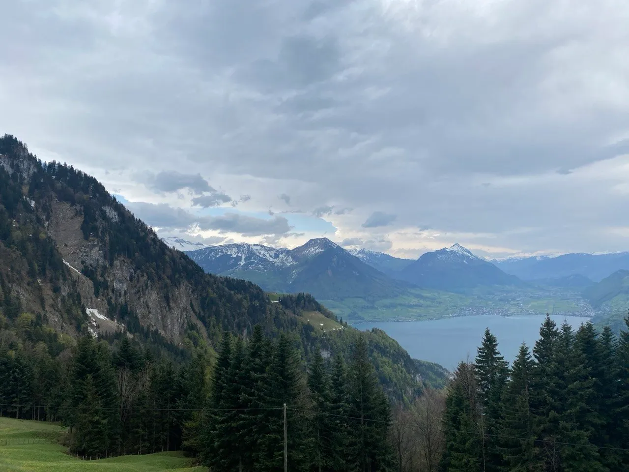 A wide view from a forested slope over the lake to a chain of snow-capped mountains under a dramatic sky.
