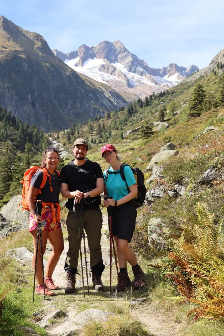 Three hikers pose together on a trail, with a snow-capped peak and glacier behind them.