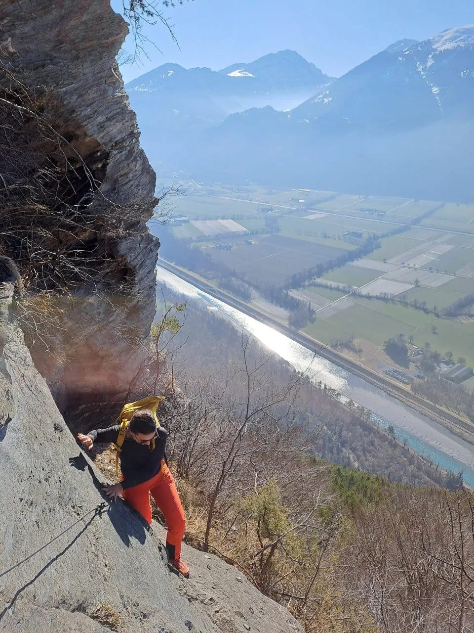 A climber descends an exposed rock ledge high above the Rhine valley, fields and river far below.
