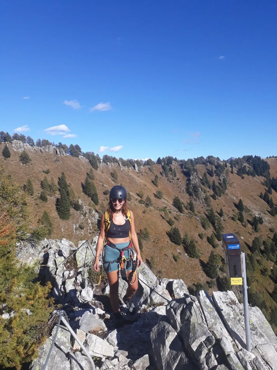 A helmeted climber stands at the exit on a rock ridge, a wide, sunny mountain crest behind her under a blue sky.