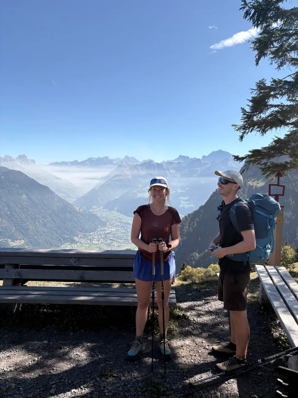 Two hikers at the Wanggrat barbecue spot, with the Reuss delta and hazy peaks behind them.