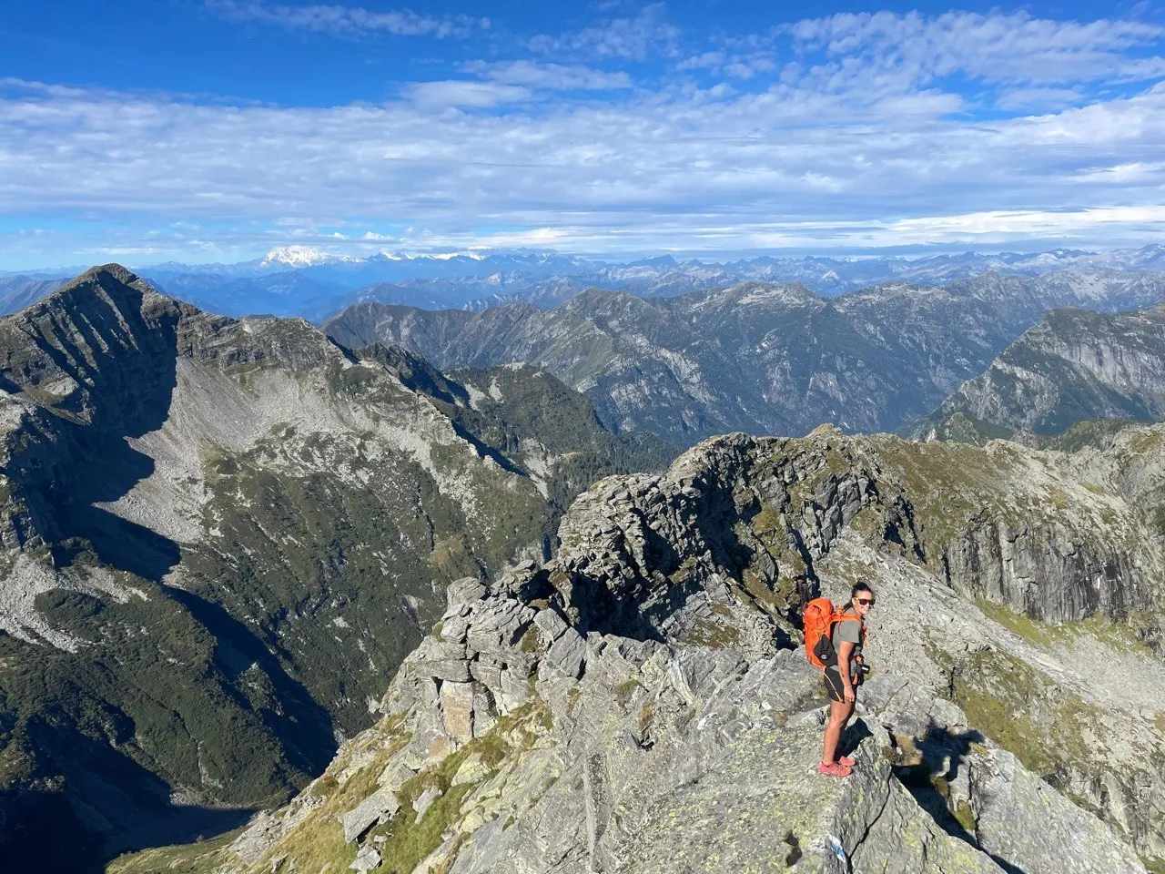 Hiker with an orange backpack standing on a rocky ridge above an endless panorama of Ticino peaks.