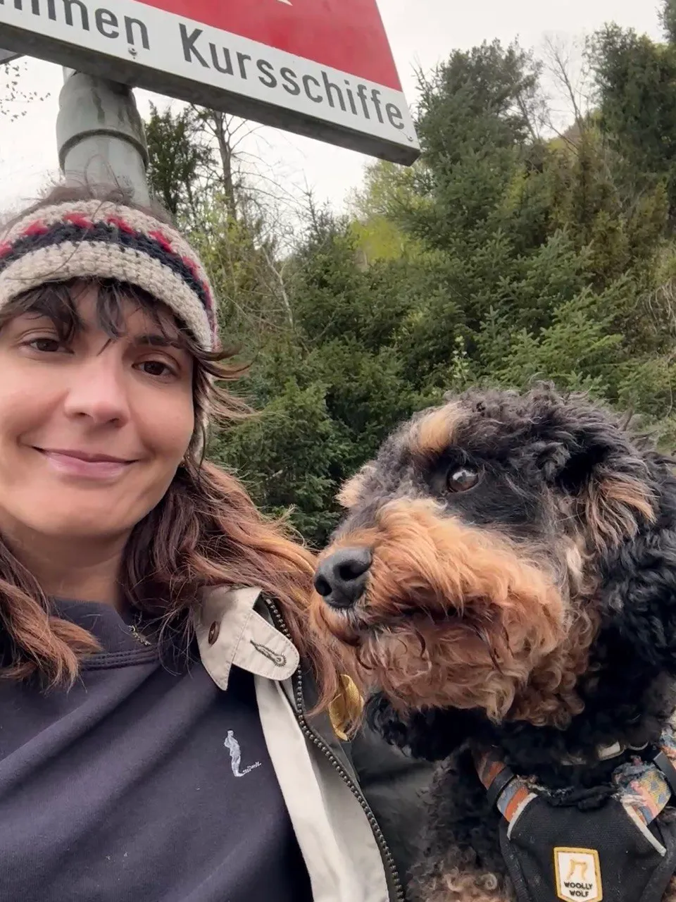 Selfie of the hiker in a knit headband with her curly dog beside a red Kursschiffe boat-stop sign.