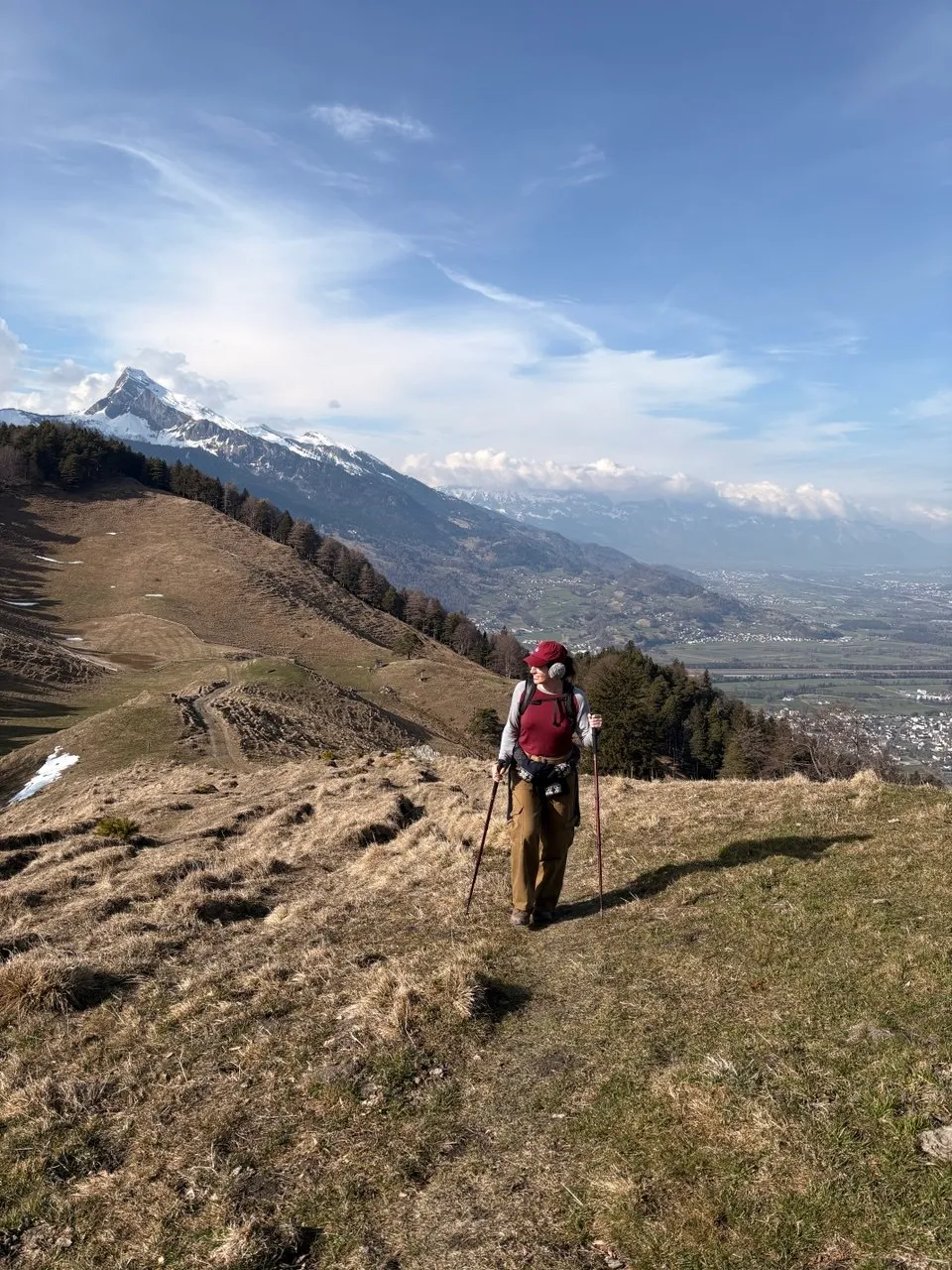 A hiker with poles and a red cap stands on a grassy ridge, a snow-dusted peak and the Rhine valley spread out far below.