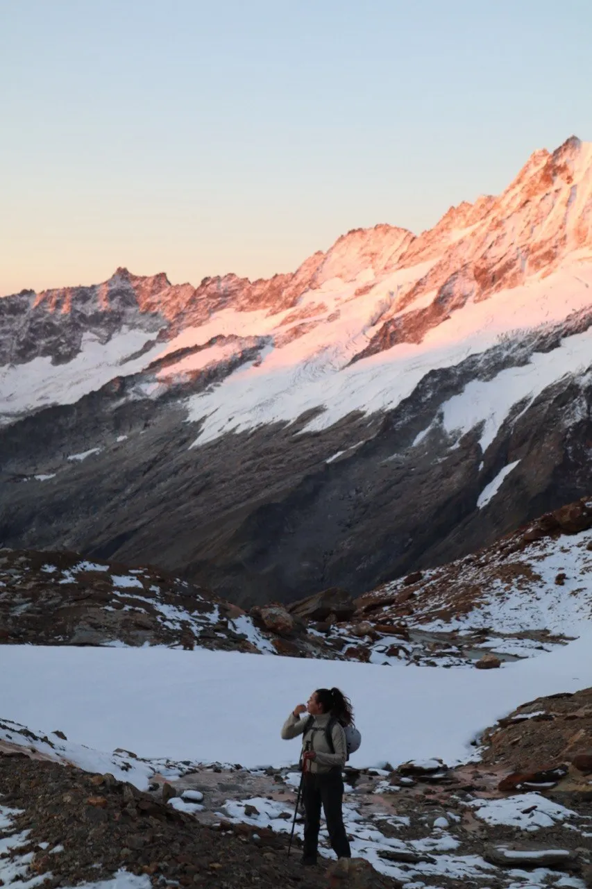 Hiker drinking from a bottle, pink-lit peaks and a snowfield behind her.