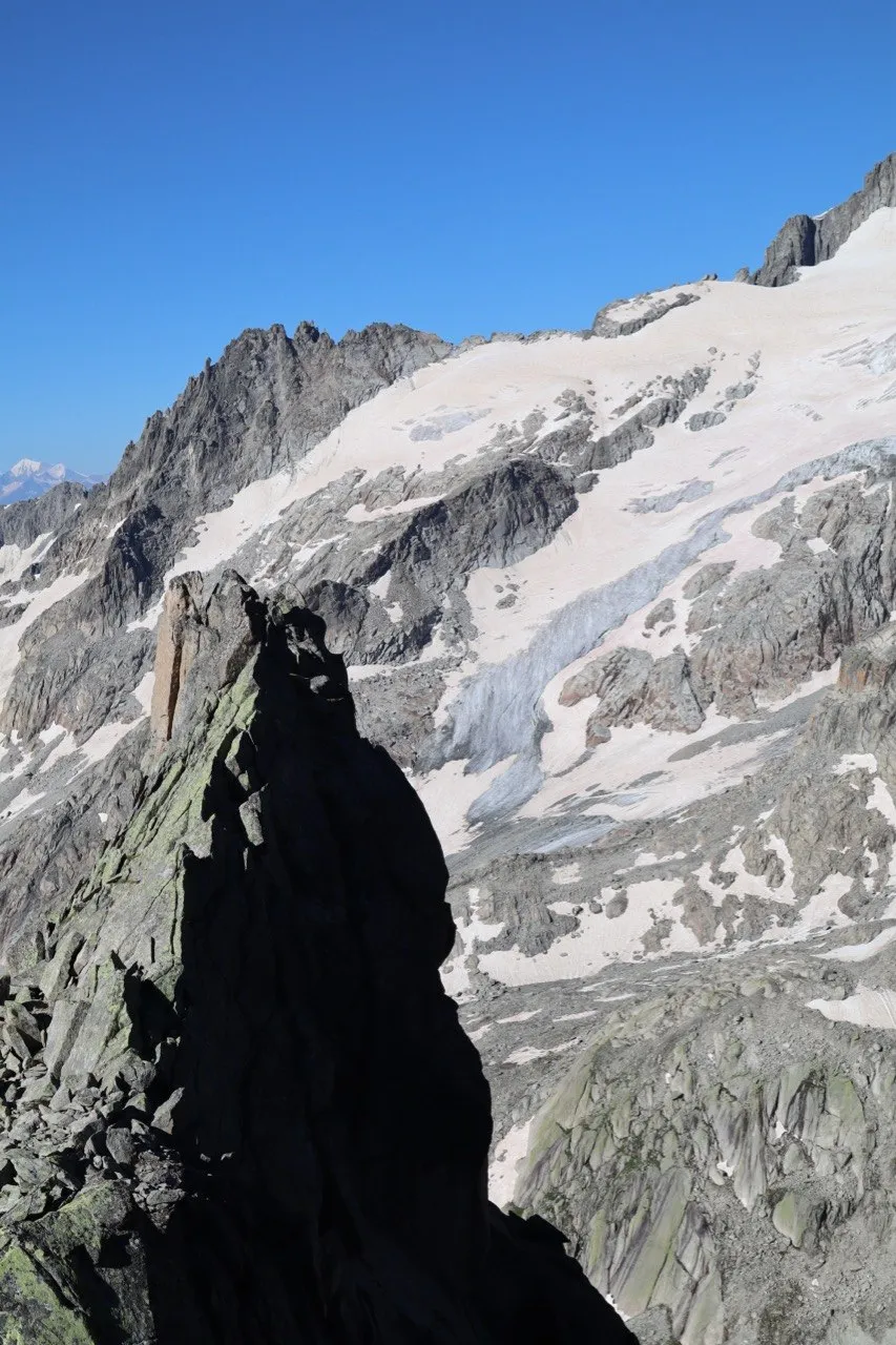A dark rock pinnacle silhouetted against a sunlit snowfield and glacier.