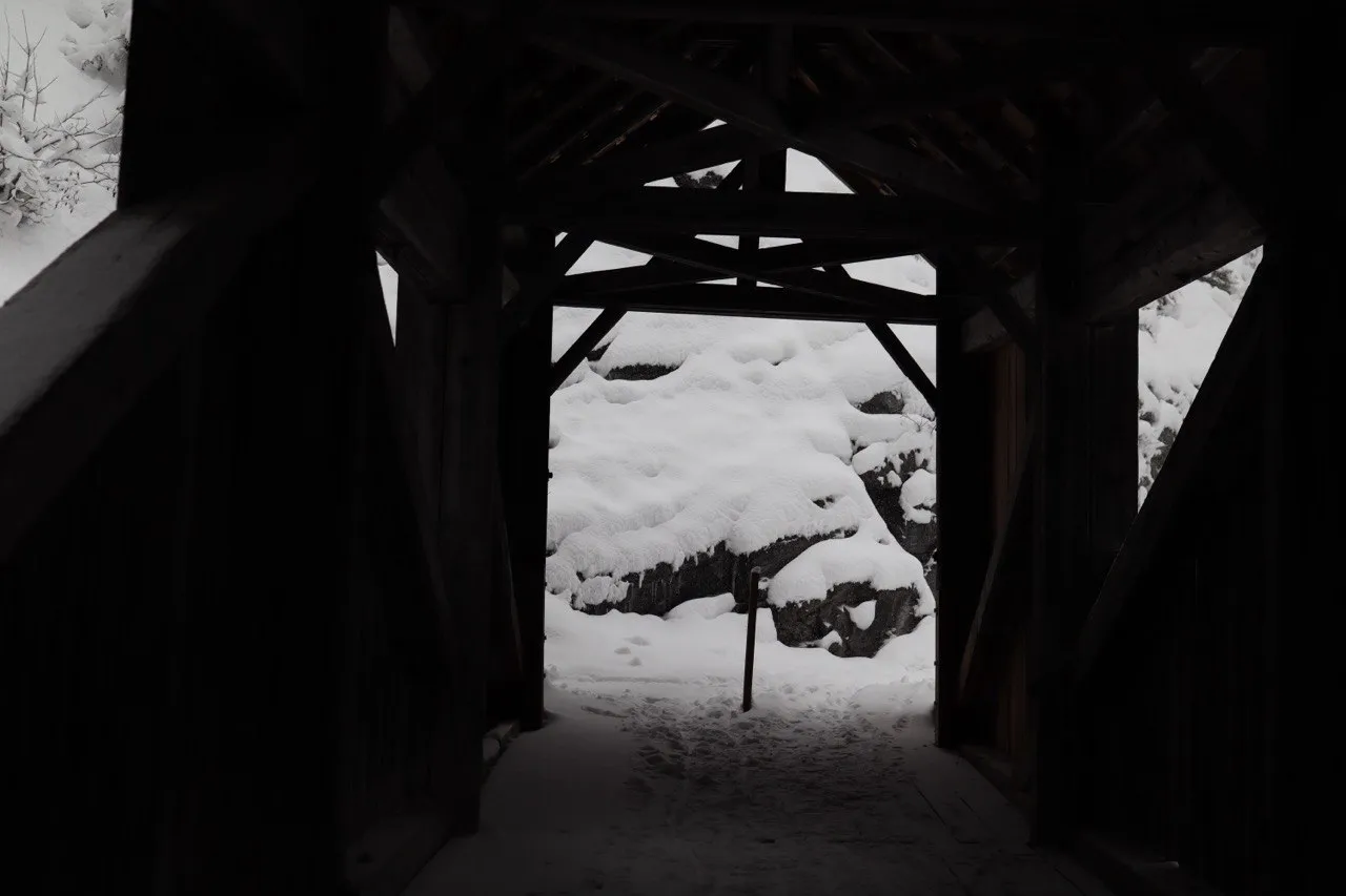 Looking out from inside a dark covered wooden bridge onto a snow-covered streambed.
