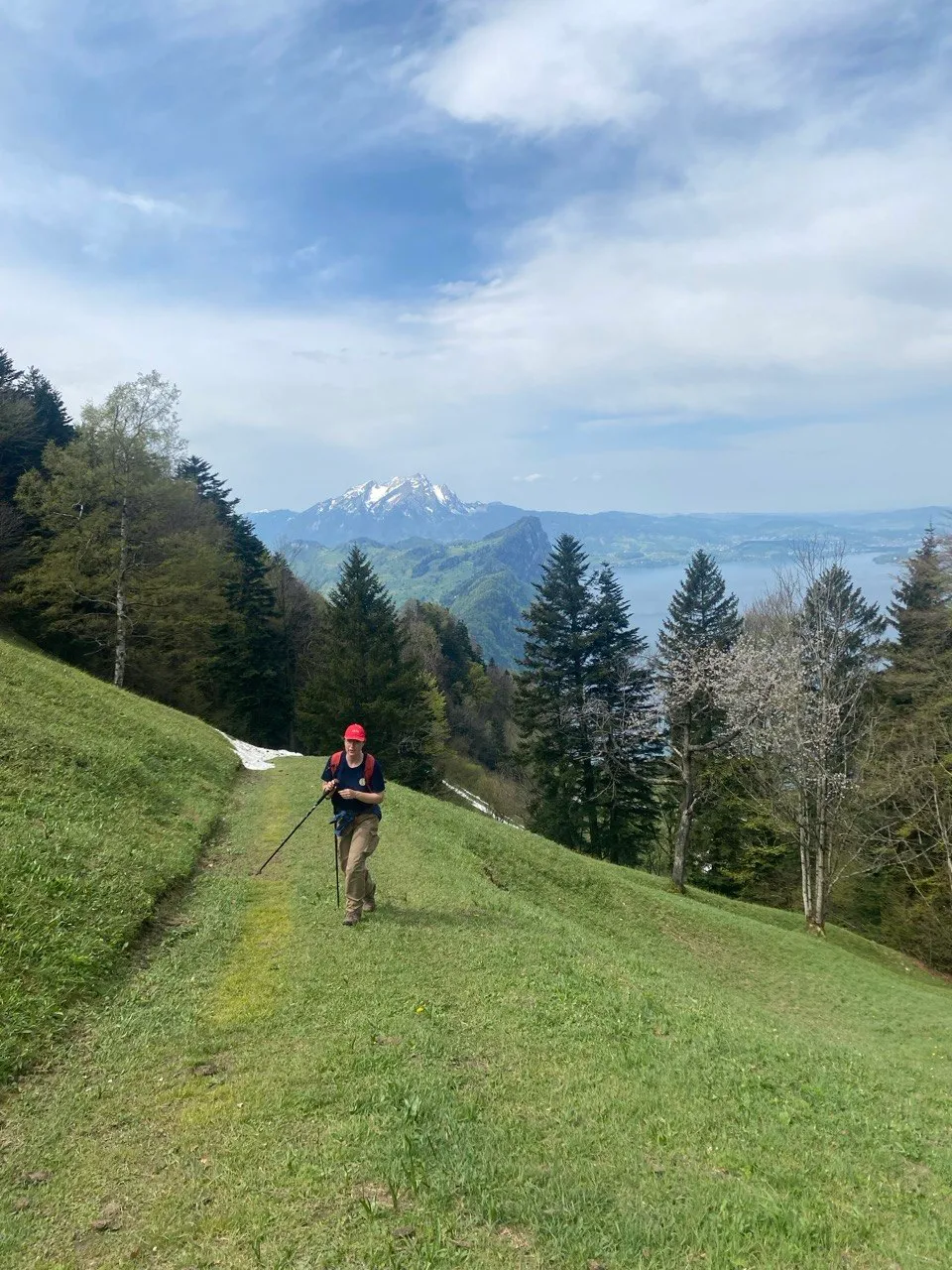 A hiker in a red cap climbs a grassy meadow, a snow-capped peak and the lake behind him.