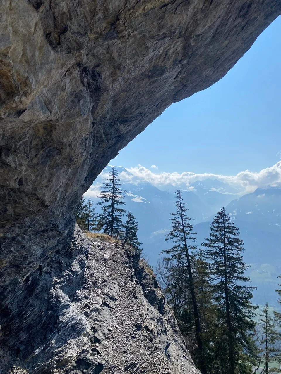 Vista bajo un llamativo saliente rocoso hacia un sendero estrecho que bordea la pared de roca, con nubes flotando sobre un amplio panorama alpino al fondo.